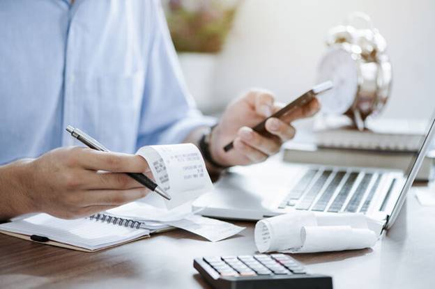 A man reviews receipts, a phone, a pen, a calculator, and a laptop are also on the desk.